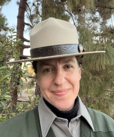 A smiling woman wearing a park ranger flat hat with pine trees in the background