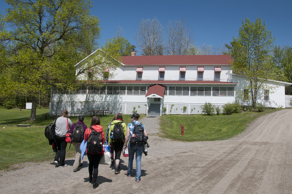 A group of adults carrying backpacks and gear approach a large, historic, white building with a red roof and a large screened porch.