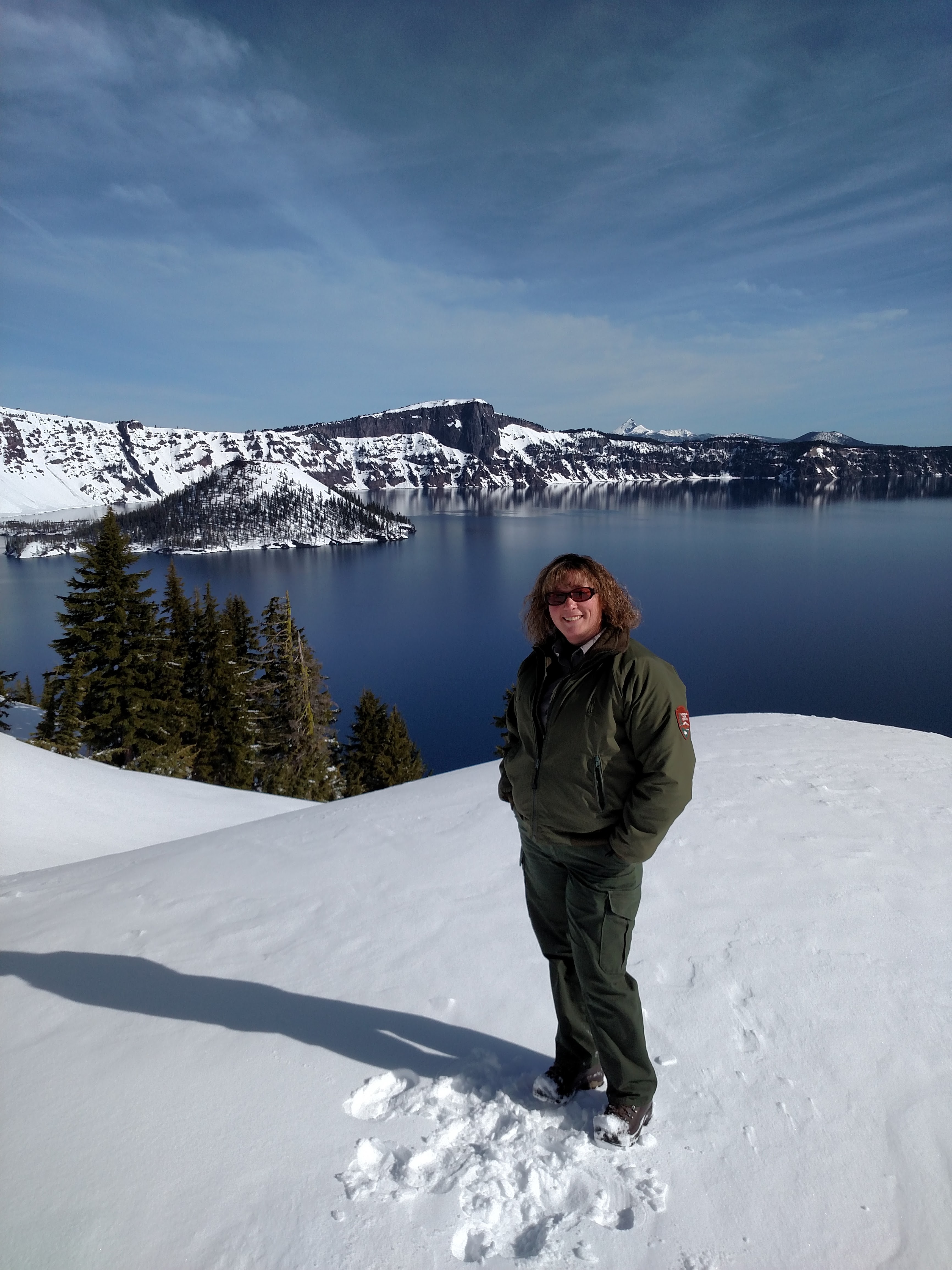 Ranger Laura standing in the snow above blue-sky water Crater Lake on a sunny day.