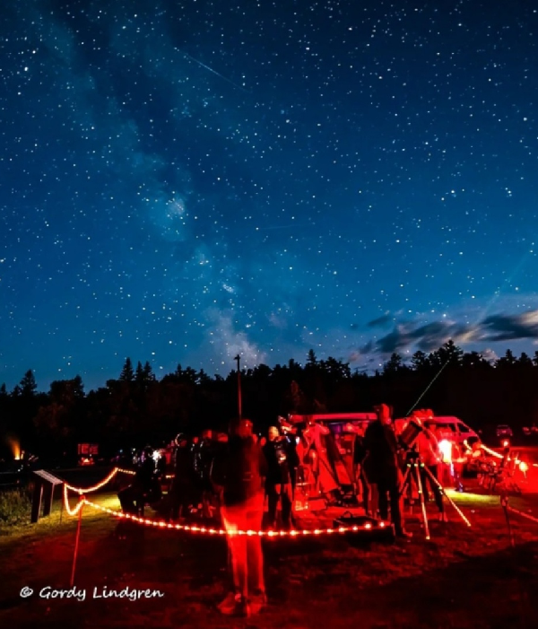 Group of people stargazing with telescopes under a clear, star-filled night sky in a forest clearing with red safety lighting illuminating pathways.