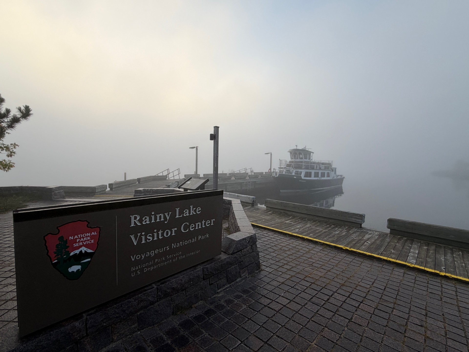 Foggy day showing the Rainy Lake Visitor Center sign with the official NPS arrowhead logo. In the background is the lake, the dock, and a tour boat
