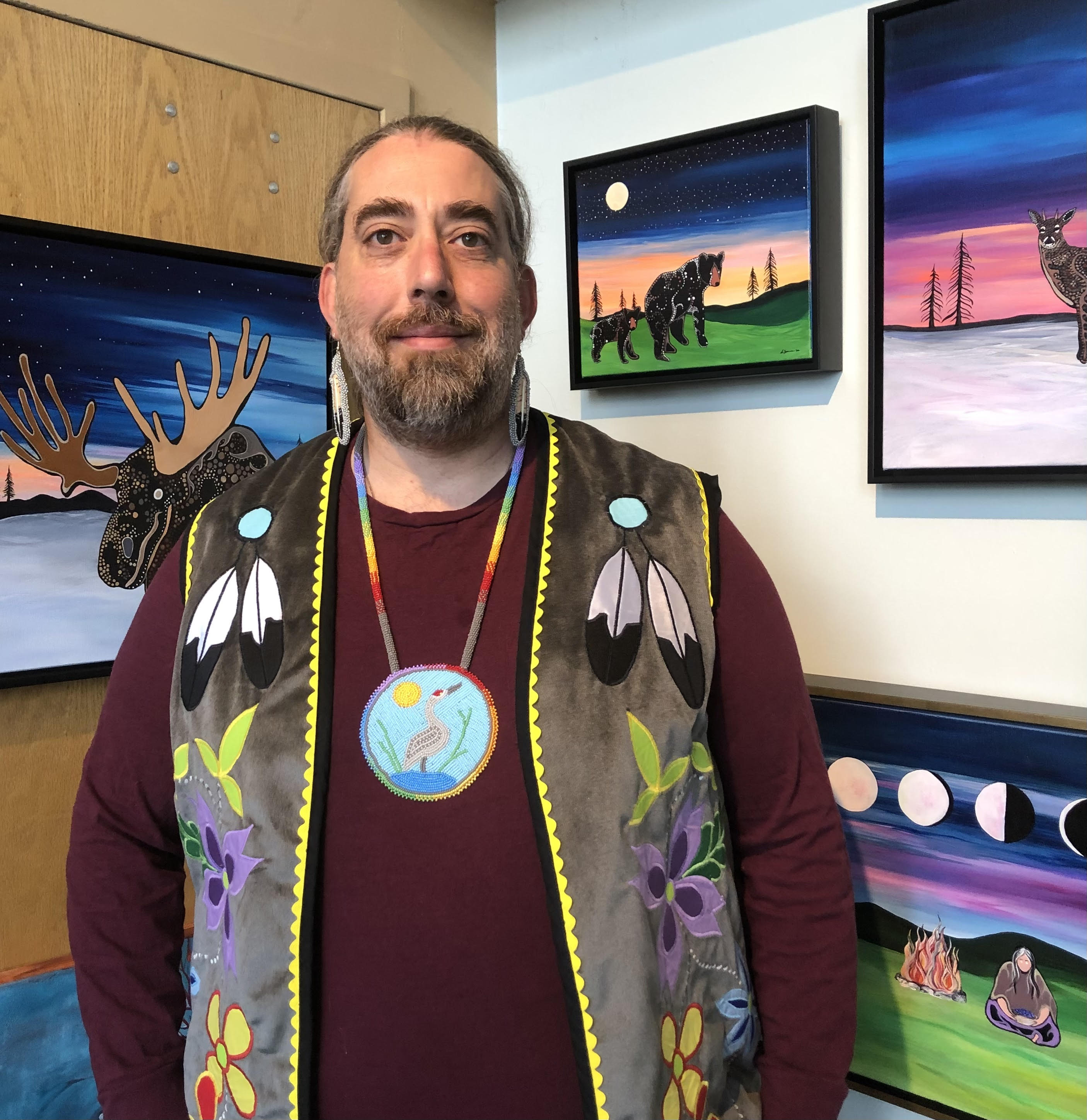 A man with a beard is wearing a colorful vest, bead necklace and red shirt stands in front of animal paintings.