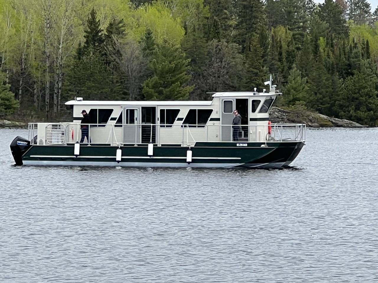 Underway view 4 green and white catamaran vessel on calm waters, green trees and rocky shore in background