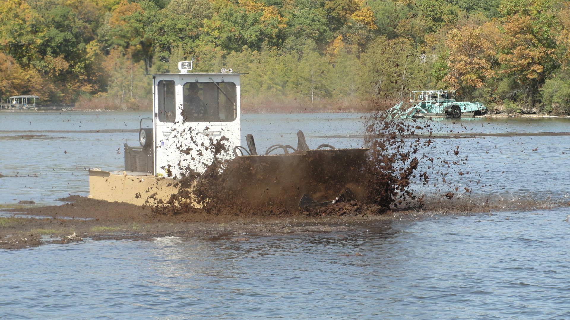 A machine on the water is shredding through brown, boggy material, sending mud flying into the air. The operator’s silhouette is visible inside the white, booth-shaped pilothouse.