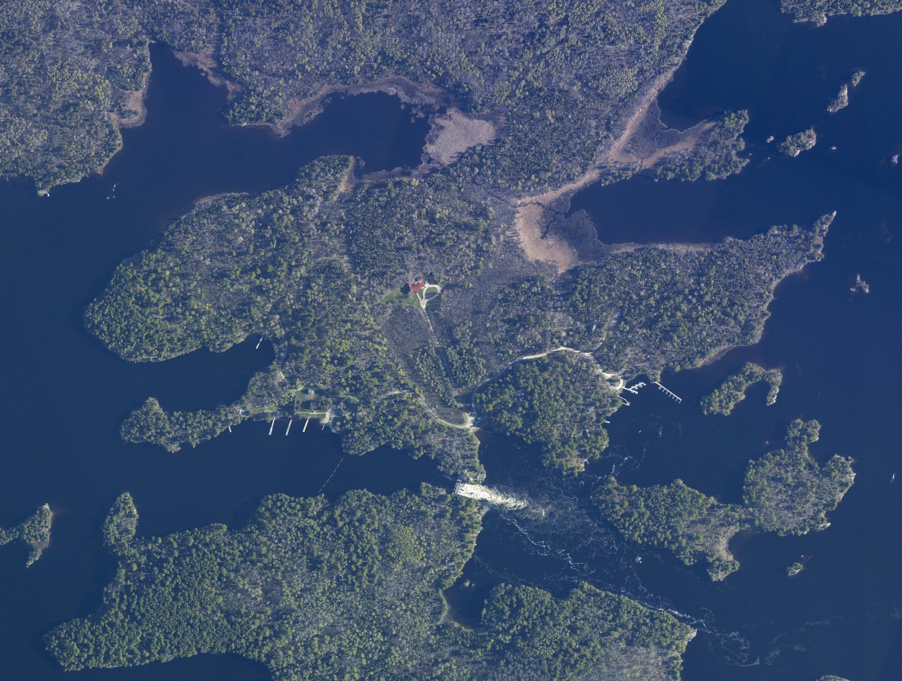 An aerial photo of the Kettle Falls area shows forested wooded areas surrounded by blue waters. Theres a cascading dam, docks, white paths, and the red roof of the hotel also visible from above.