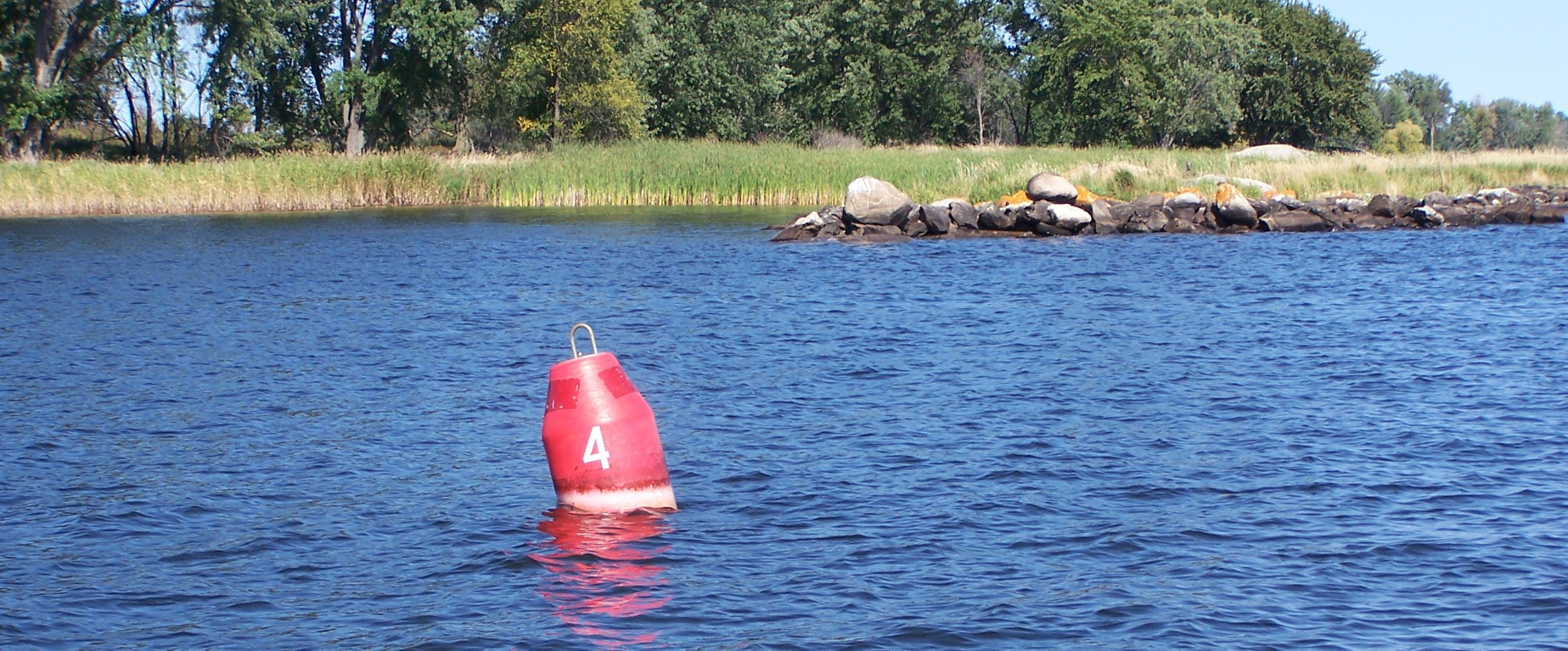VOYA_Red_Nun_Ram_Island_5 A red buoy in a blue lake with trees in the background on the edge of the water.