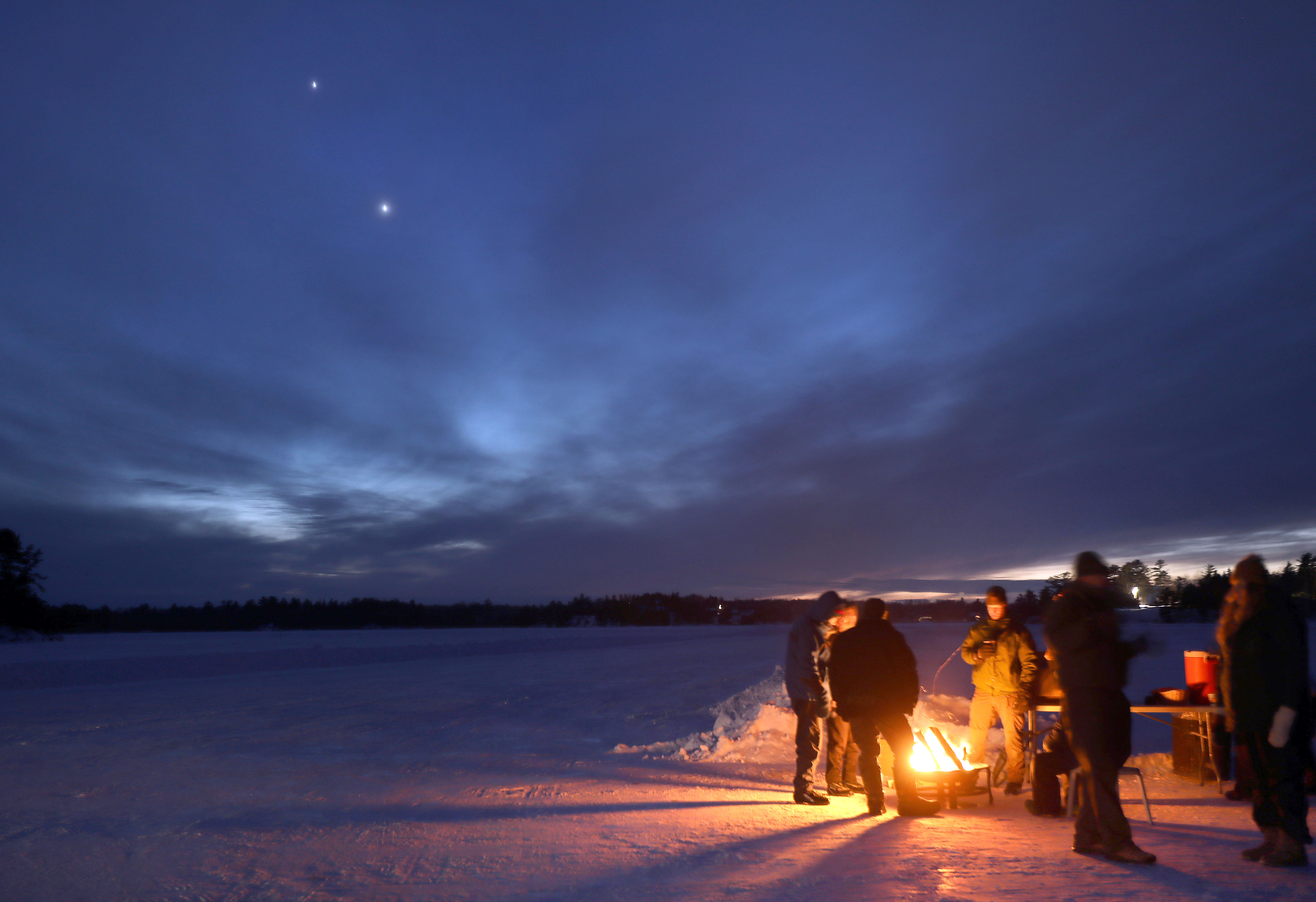 5 people gathered around a small fire on a frozen lake.  The sky is dark, but two bright planets are visable low on the horizon.  Jupiter is just below venus.