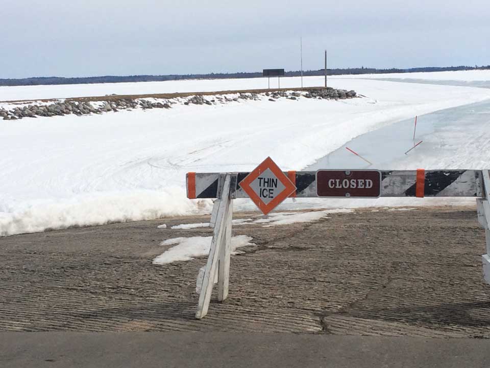 Ice Road Closed Road barricade with a closed sign and thing ice sign blocking access to a frozen lake surface in background.