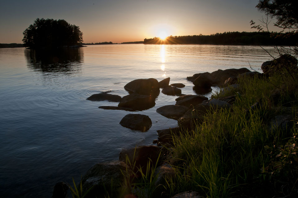 Sun is setting over a tree line, with a reflection in the water. Island located in background with a rocky shoreline in the foreground.