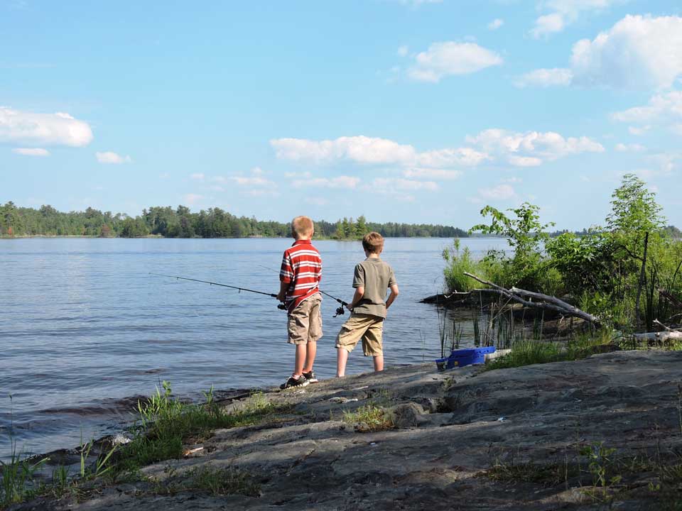 Two young boys fishing from a rocky shoreline, holding fishing rods, trees and open water in background.