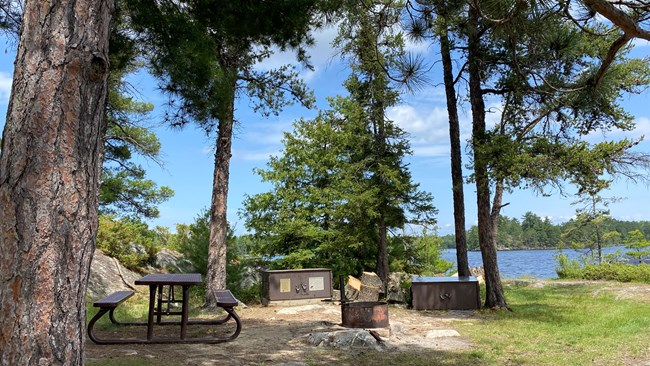 A fire ring, table, and two metal food storage lockers sit on an island shoreline