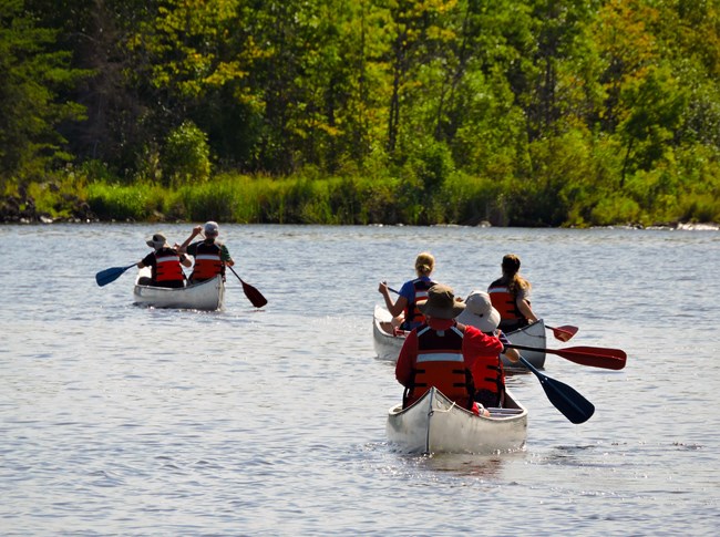 Six adults paddle three metal canoes across the calm waters of a scenic lake, headed for the opposite shore.