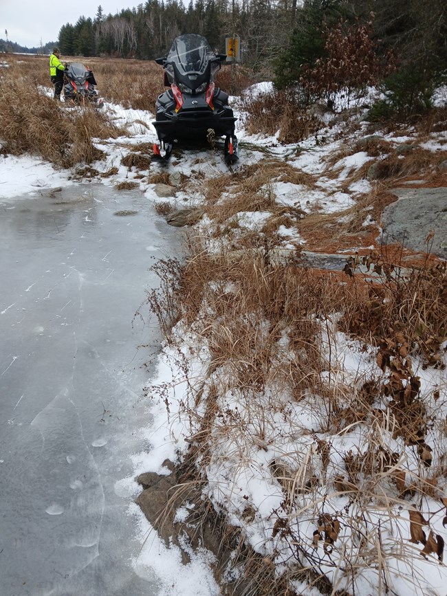 Two snowmobiles and one rider can be seen on a trail that has patchy snow cover and ice