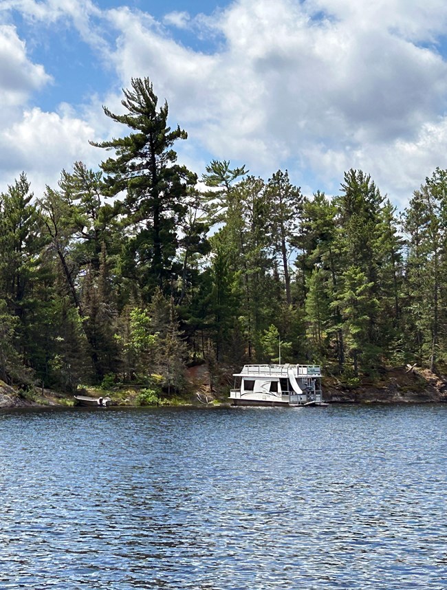 A boxy, white, double-decker houseboat with a stern waterslide floats on the shore of a scenic lake, with its bow ashore, surrounded by pine trees.