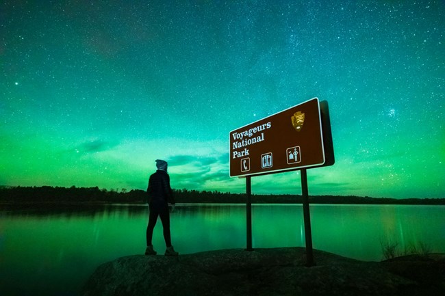 Photograph of person standing next to Voyageurs Park sign at night under Norther Lights.