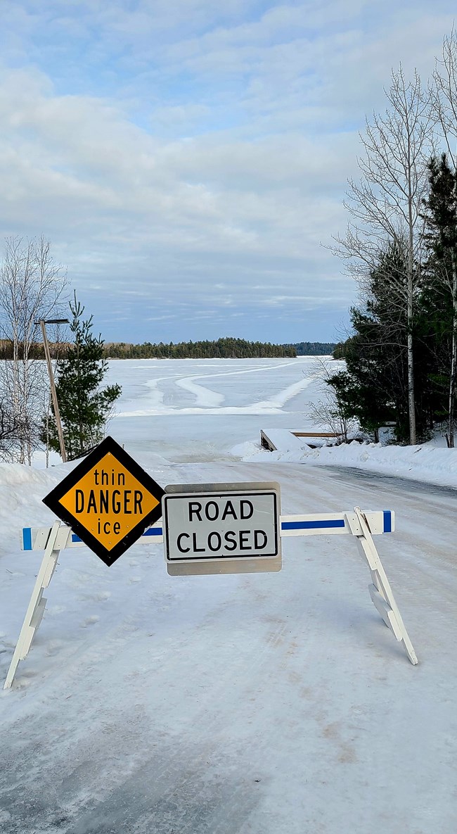 A road closed and thin ice sign in front of a frozen ice road.  trees are in the foreground.