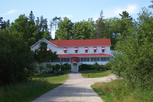 A red roofed hotel is surrounded by green trees with a path leading to the front steps. The hotel is two stories with windows and white siding.