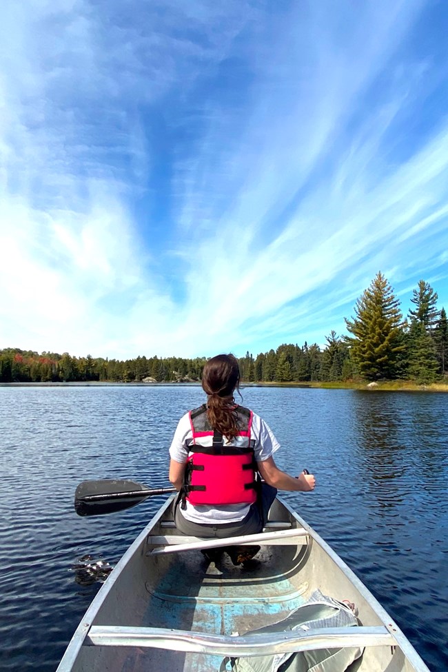 A woman sits in the bow of a metal canoe, holding a paddle, facing away towards the tree-filled shoreline of a scenic lake.
