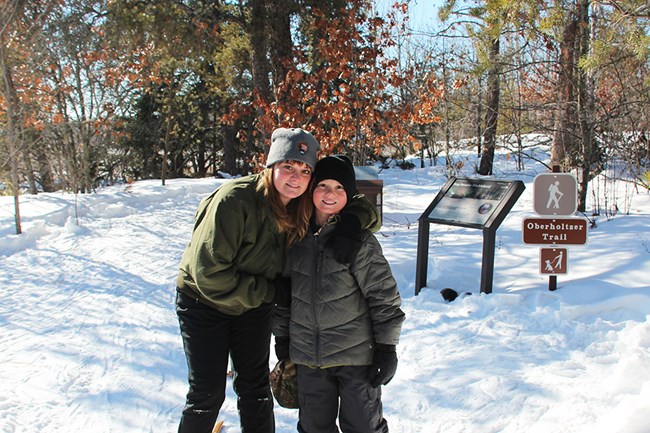 A mother puts her arm around her son's shoulders and both smile while standing at the head of a sunny, snow-packed trail.