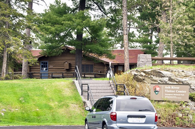 A blue van is parked in front of a log building. Near the van, a sign reads "Ash River Visitor Center."