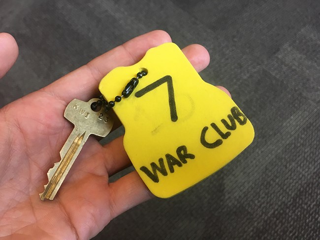 Key for a Backcountry Canoe A hand holds out a brass key that is linked by a small chain to a large, yellow float labeled "7 War Club."