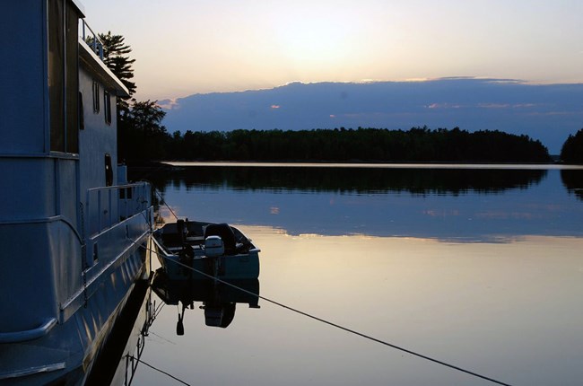 A large houseboat and a small fishing boat are seen in profile, moored side by side and facing a dark sunset on the horizon.