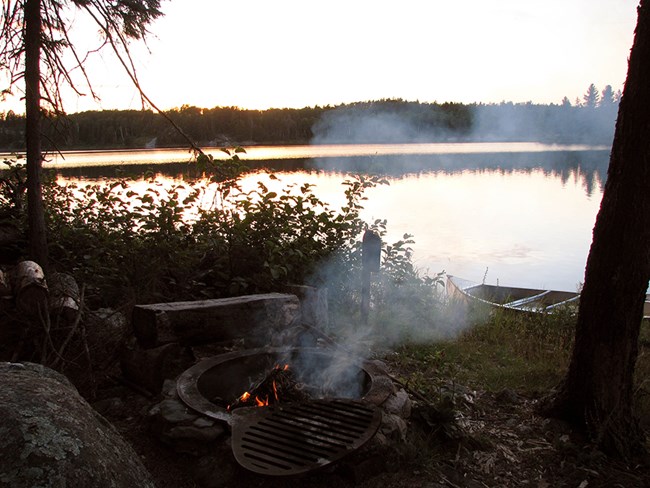A small campfire burns in a metal fire ring on the shore of a scenic lake at dusk. Behind the campfire, a canoe floats on the shore.