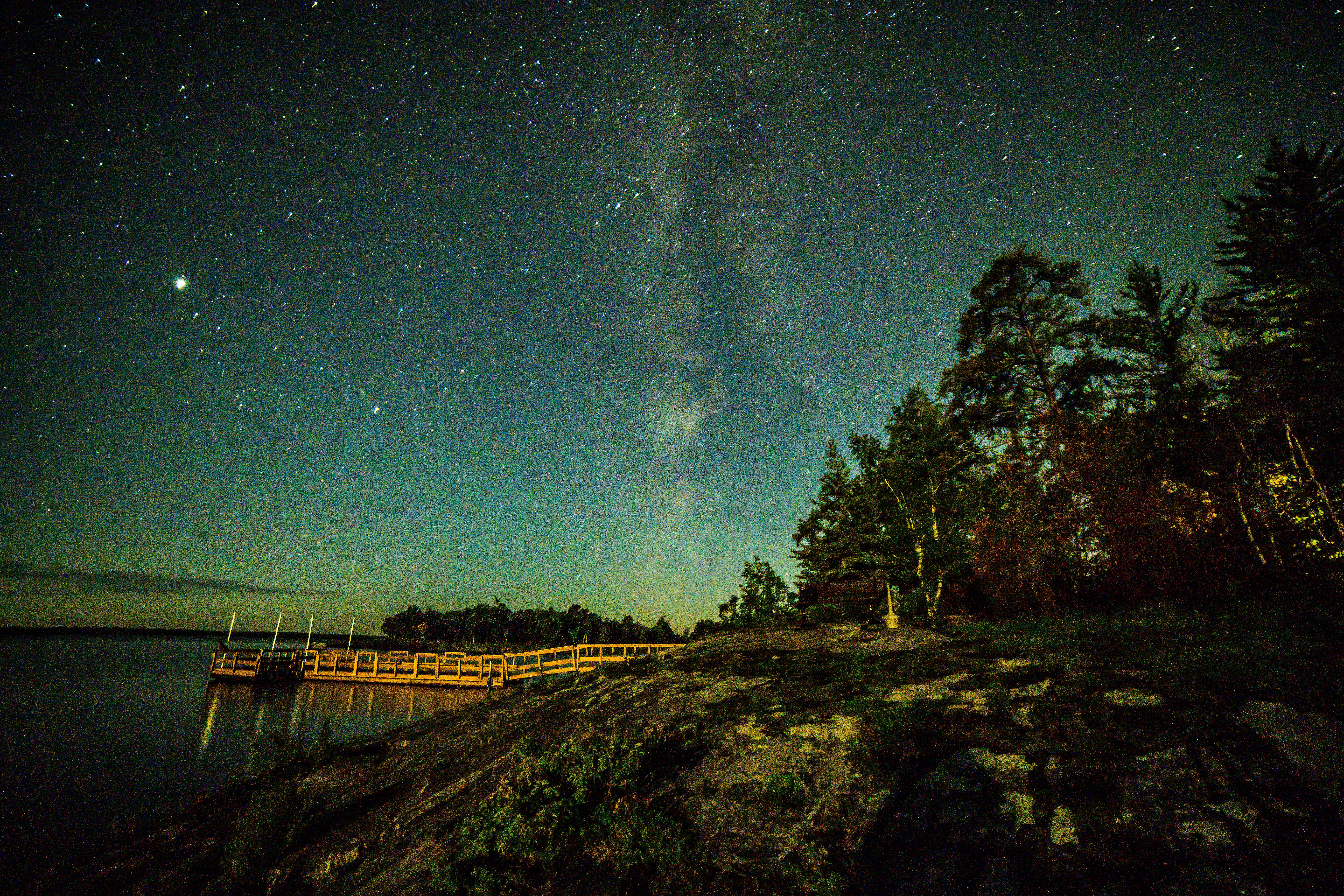 Millions of stars shine vividly in a line-like cluster over the shoreline of a scenic lake, which reflects their image.