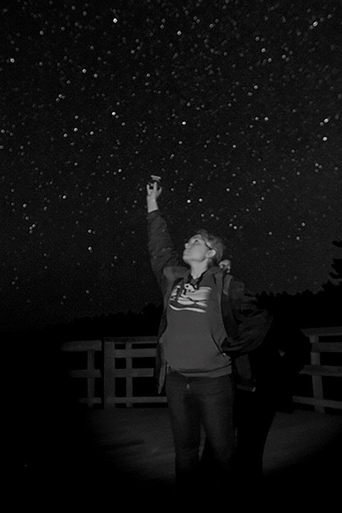 A woman gazes up at a star-filled dark sky, holding a small, round metal piece of equipment aloft over her head. Silhouettes of trees and a wooden deck in the background.