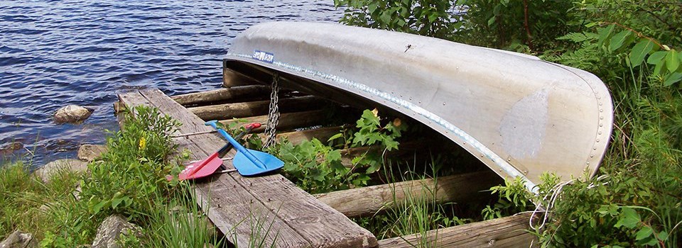 A canoe sits upside down on a wooden rack on the shores of a scenic lake, secured to the rack by a chain. Two paddles lay crossed next to the canoe.