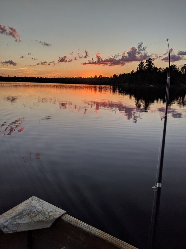 The orange glow of a sunset reflects on the calm waters of a scenic lake. A black fishing pole in the bow of a metal boat faces the dimming horizon.