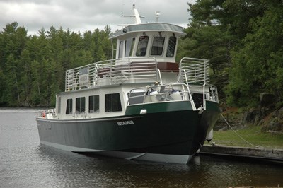 A tour boat docked on water at a wooden boat launch with green grass on the shore and trees in the background. The sky is cloudy. The tour boat is 2 tiered with windows and a captain's quarters. The bottom of the boat is black and the top is white.