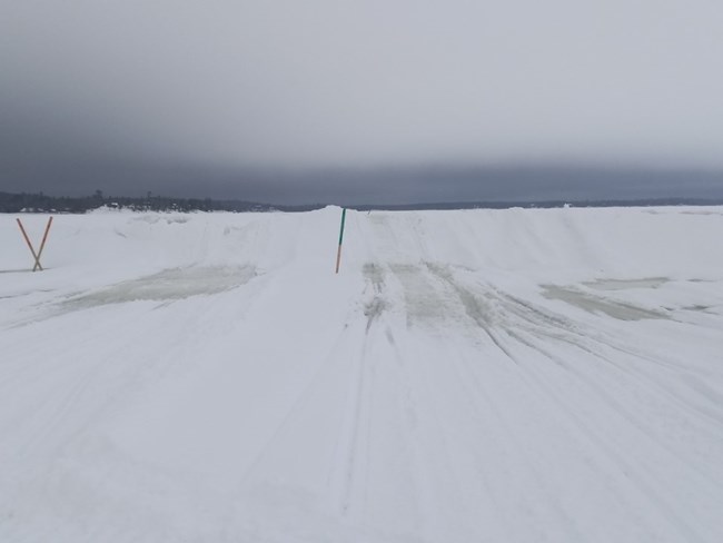 A large, sheer hill of ice rises suddenly from a flat snowmobile trail, marked by two orange hazard stakes.