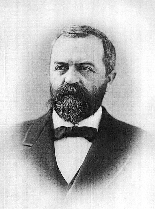 A portrait of a man with a full beard wearing a black tuxedo and white dress shirt.