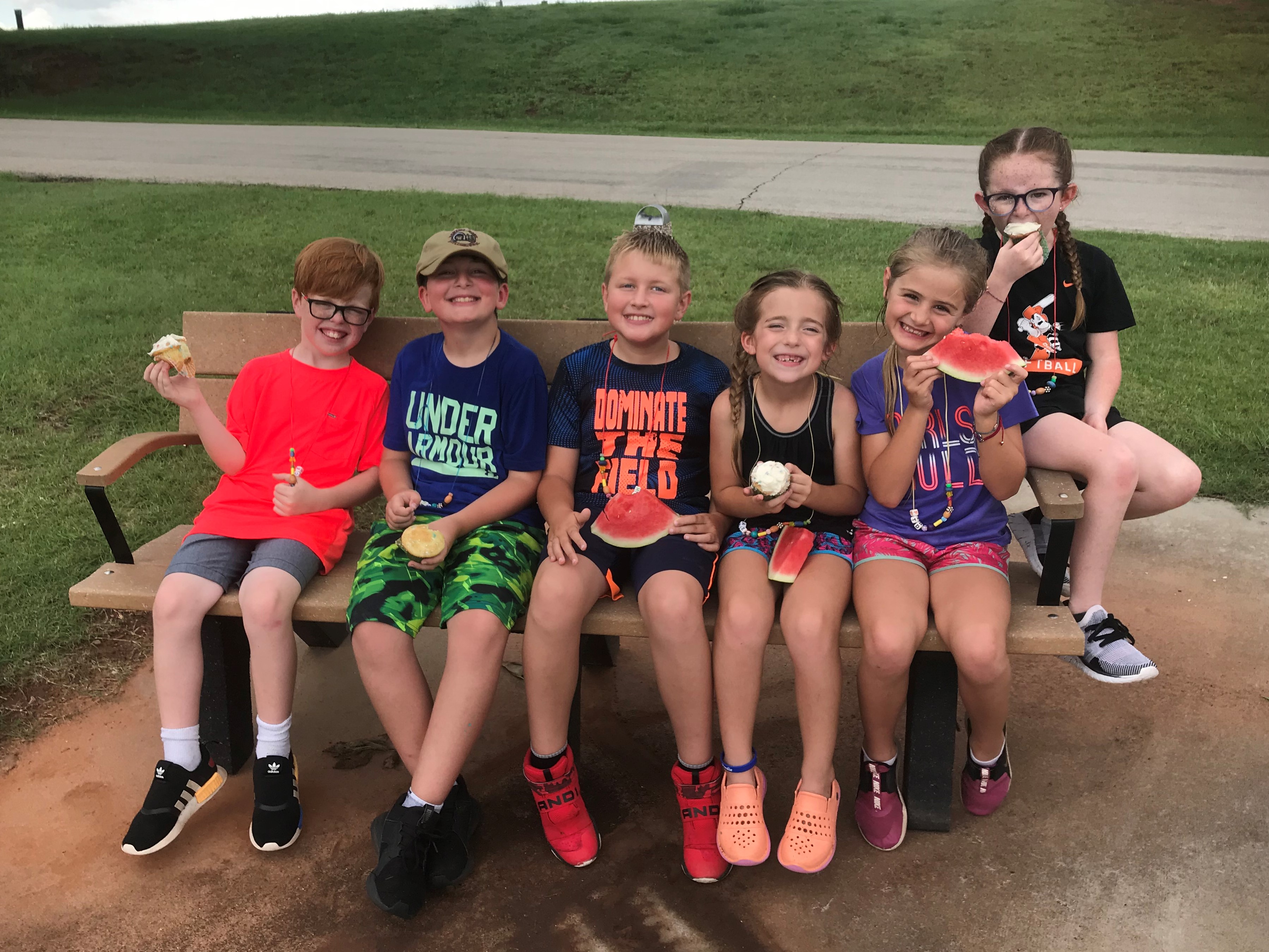 Kids sitting on a bench eating cupcakes and watermelon.