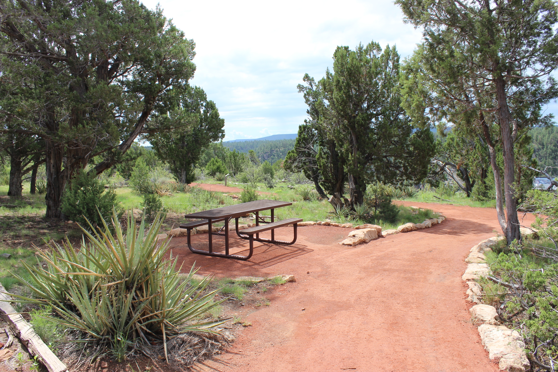 A visitor center sits across the canyon at cliffs edge surrounded by pine trees. The San Francisco peaks mountain range rises out of the horizon, dwarfing the canyon in comparison.