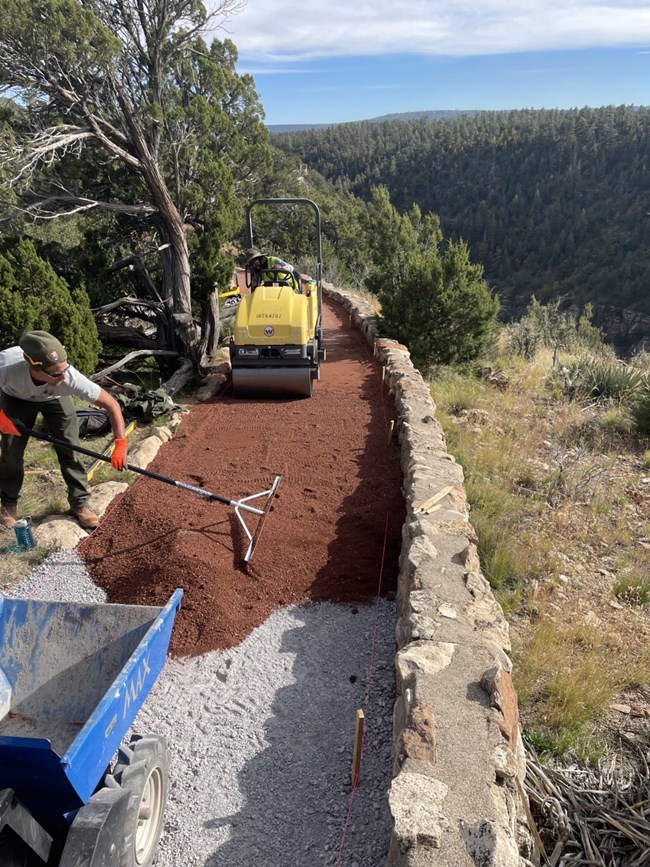A park ranger rakes material over a trail while another park ranger drives a compator over the materal