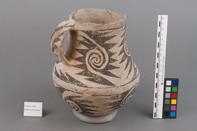 Ancient black-on-white pitcher excavated at Walnut Canyon National Monument. An ancient ceramic pitcher made of white clay with angular, black designs painted onto it. The pitcher is in a sterile setting with a label identifying it as "WACA 964, MNA # Unknown." Beside it are a series of color swatches and a ruler.
