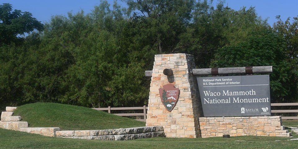 Brown Waco Mammoth National Monument sign with green grass, trees, and blue sky.
