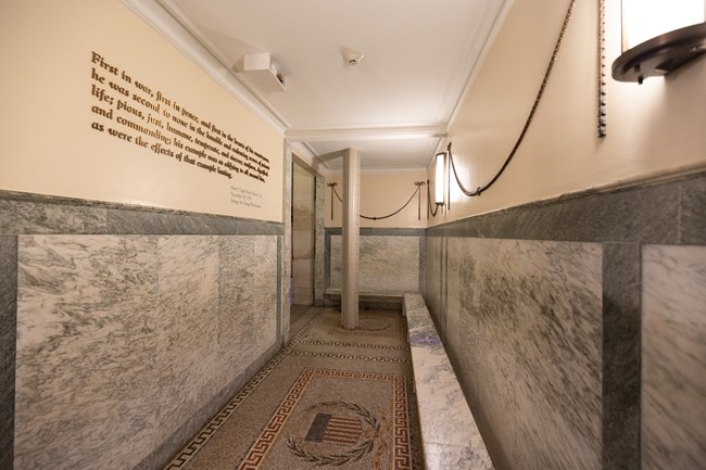 A bench lines the walls in the waiting area to ride the elevator to the observation floor. A quote is seen on the left wall and a pole embedded from the ceiling to the ground stands in the background.