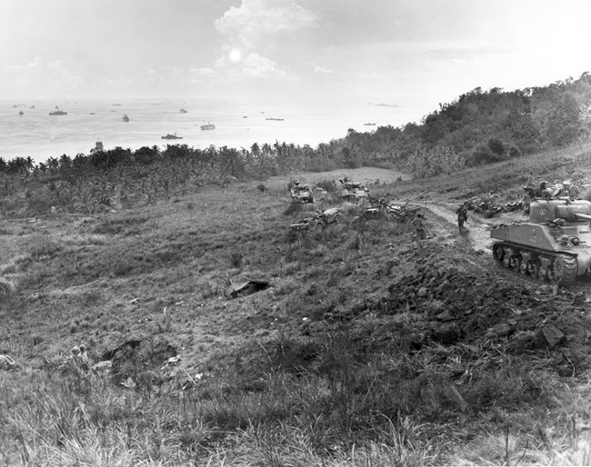 Tanks chug their way up a hill on a dirt road. In the background, battleships float in a bay.