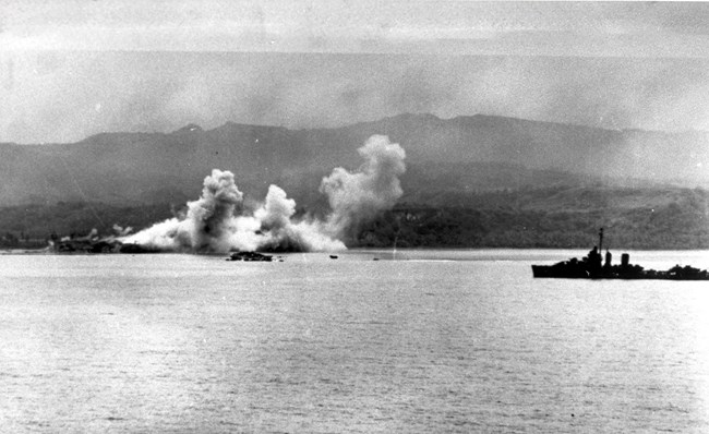 A battleship floats on the ocean in front of an island. In the background is a large explosion.