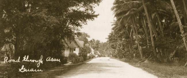 Aged photo with caption "Road through Asan, Guam"; scene captures a wide straight road with vegetation and trees on either side. In the distance, on the left side, resides a few homes with thatched roofs.