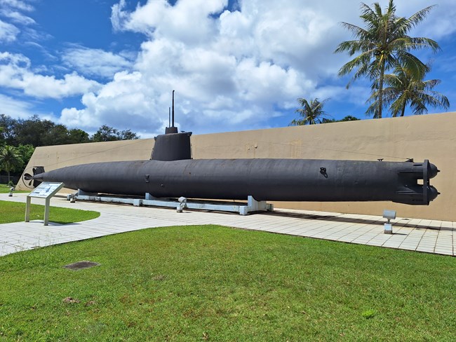 A large black submarine with a pericope rests on a concrete plinth in front of a concrete wall.
