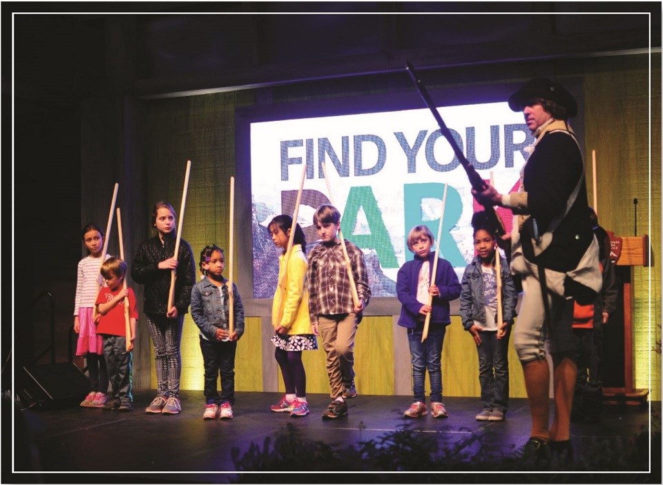 Eight children standing in a line holding replica muskets on a stage in front of a sign reading "Find Your Park". A man in period clothes holds a replica musket while standing in front of a podium.
