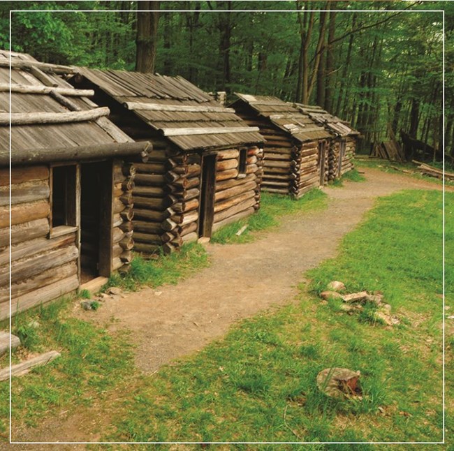 Four primitive log cabins stand in a row next to a dirt trail in front of a dense canopy of tall green-leafed trees. Each cabin has one open windown without a covering and an open doorway without a door.