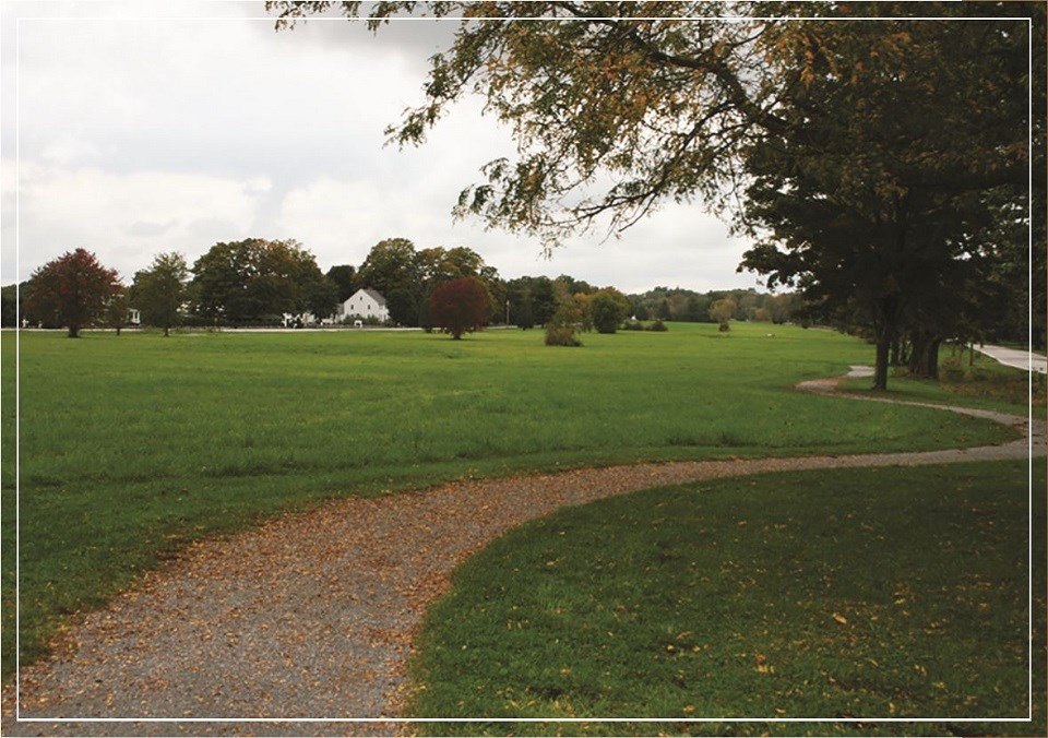 A leaf-covered gravel trail winds past trees at the edge of a green grass field. A white 2.5-story house surrounded by trees sits at the edge of the far side of the field.
