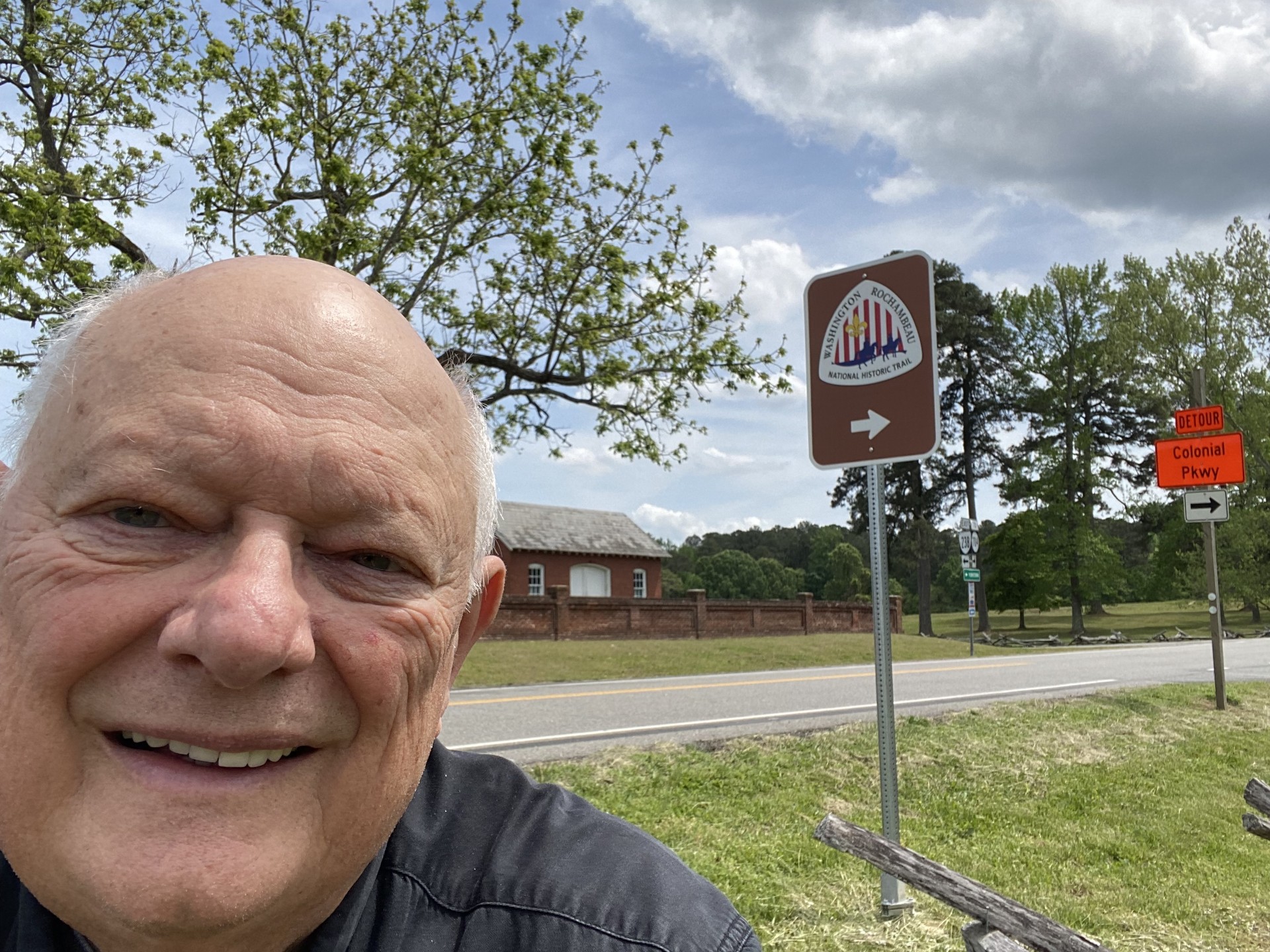 Trail enthusiast Frank Kos poses by a trail marker at Colonial National Historical Park in part of the Washington-Rochambeau route. A bald man smiles in a selfie by a trail sign