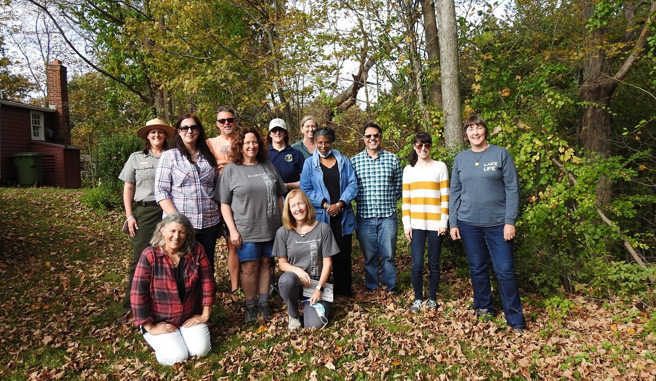 A group of people stand in a meadow with fall trees behind them.