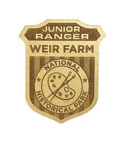 A wooden junior ranger badge with two paint brushes and a palette in the middle and the words, "Junior Ranger Weir Farm National Historical Park".